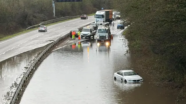 A car in flood water on a large road. There are cars and vans stopped just before the flood water starts and people in high visibility clothing.