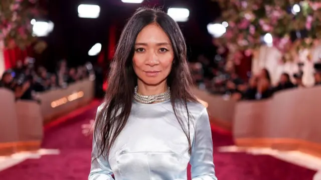 Chloe Zhao wears an ice blue silk outfit with a diamond choker-style necklace - she has small plaits in her long dark hair - she is standing on a red carpet with photographers just seen in the background