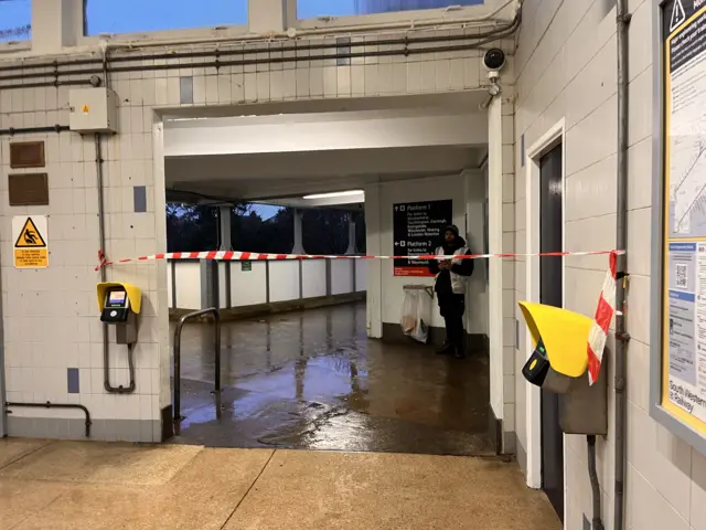 Red and white tape blocking the station entrance at Pokesdown. One worker can be seen behind it. The station floo r is flooded.