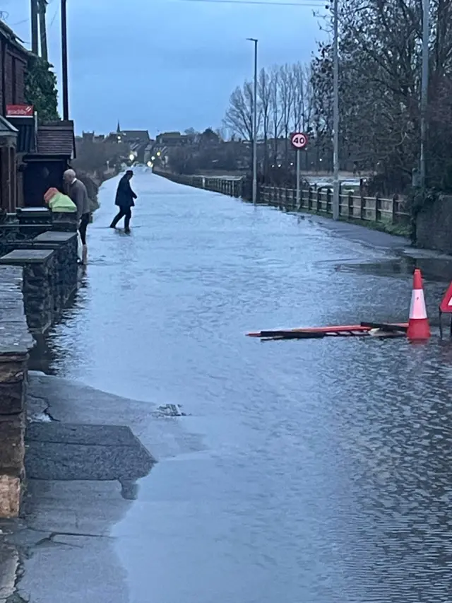 A flooded Stoborough end, leading to Wareham High Street, near Ridge in Dorset. Some people are working on the sides of the road. Cones and signs can be seen in the water.