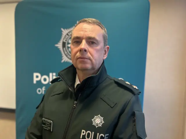 Man standing in PSNI uniform in front of a tall PSNI banner. His uniform is green with a PSNI logo and he has short grey hair