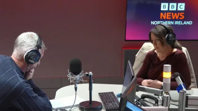 A man and a woman in a radio studio - the man on the left has his chin in his hand and is looking down at a piece of paper. The woman on the right is waiting for a question