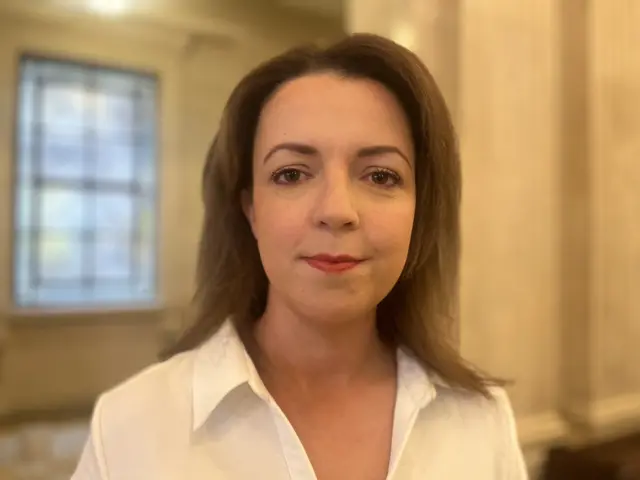 Liz Kimmins, a woman with straight, dark, shoulder-length hair, looks to the camera in Stormont. She is wearing a white shirt with an open collar.