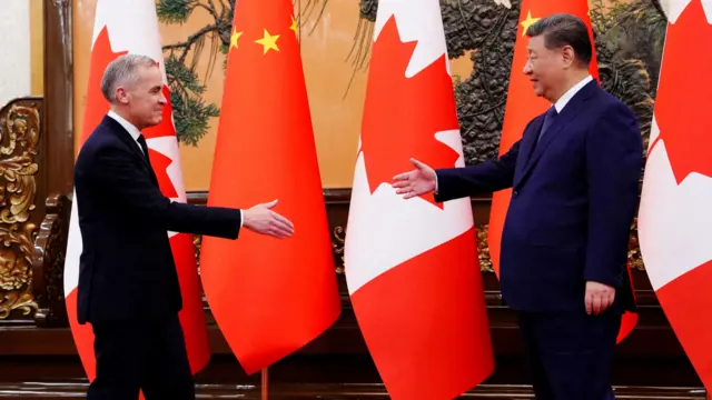 Canadian PM Mark Carney extends his arm to shake hands with Chinese leader Xi Jinping, both wearing suits and standing in front of large flags of their countries