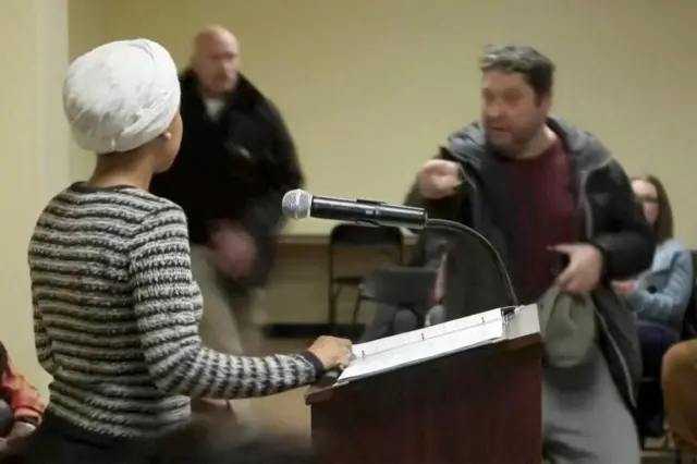 A man in a dark jacket and maroon sweater points at Rep. Ilhan Omar