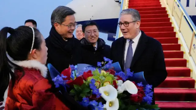 starmer is presented with a bouquet of flowers by a woman in a red coat. he stands in front of the red carpeted steps of his plane.