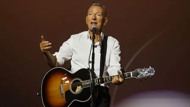 Bruce Springsteen performs onstage with a guitar. He is wearing a long sleeve white shirt and black pants
