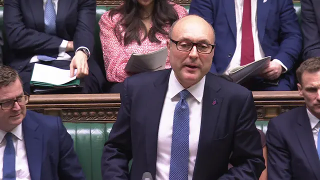 Andrew Griffith is speaking at deputy PMQs wearing a blue suit and blue tie. He is wearing glasses and is surrounded by conservative MPs on the benches