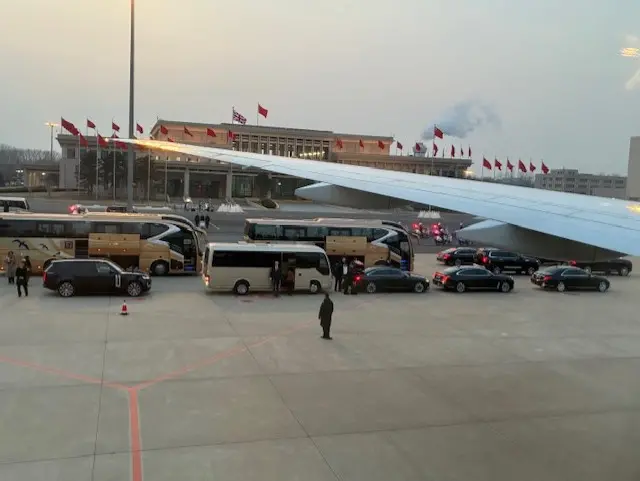 a line of buses and black vehicles line the tarmac in beijing as starmer arrives. the plane's wing can be seen out the window