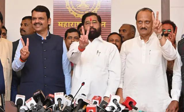 Ajit Pawar (right) with Devendra Fadnavis (left) and Shiv Sena leader Eknath Shinde (centre) holding up victory signs. Pawar and Shinder wear white while Fadnavis is in a light blue shirt with a dark blue vest