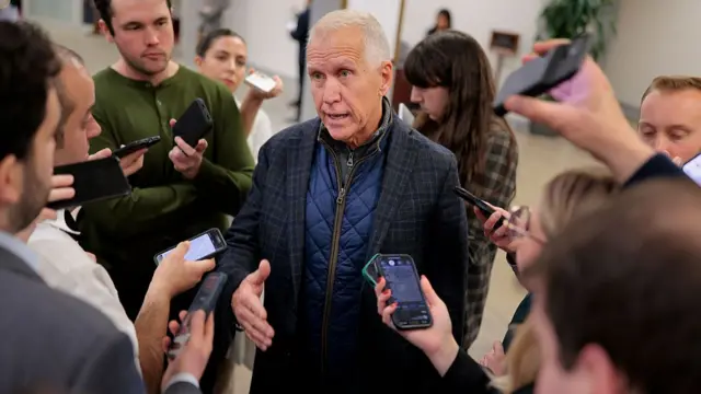 Thom Tillis speaks to reporters. He wears a checkered suit jacket with two zip-up coats layered underneath