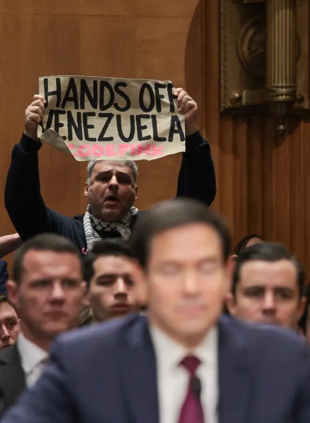 Rubio in Senate, behind him a man holds a sign saying 'hands off venezuela'.