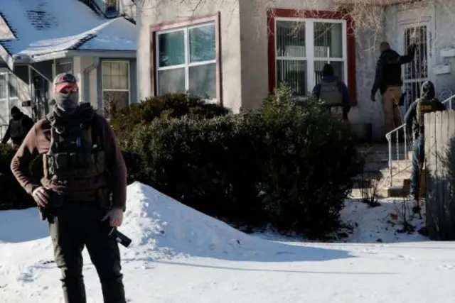 Immigration agents in tactical gear stand outside a home in a snow covered garden while one of them knocks on the door