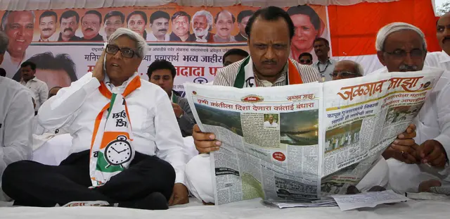 Ajit Pawar seen sitting on a stage, holding open a newspaper with election banners in the background
