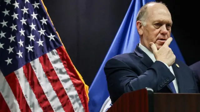 Tom Homan standing at a lectern holding his right hand to his face. Behind him is a US flag