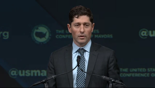 Minneapolis mayor Jacob Frey speaking. He's standing in front of two thin black mics in a dark suit, light blue shirt and striped tie
