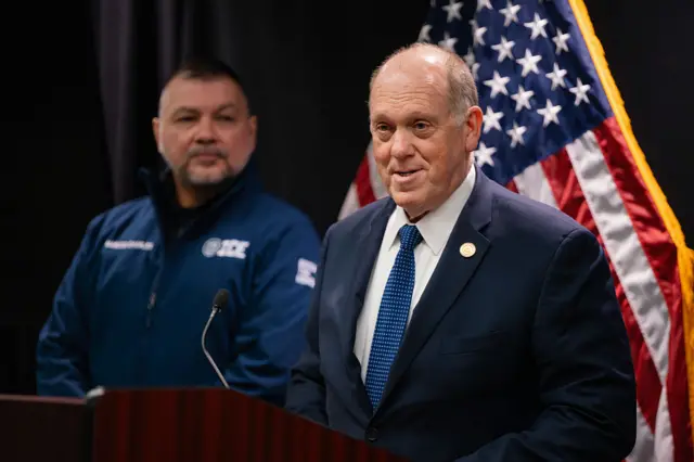 Mid-shot of border tsar Tom Homan during a press conference in Minneapolis. He's standing at a wooden lectern in a dark suit, white shirt and blue tie. Behind him, to his right, is an ICE officer in a blue jacket standing next to a US flag (R)