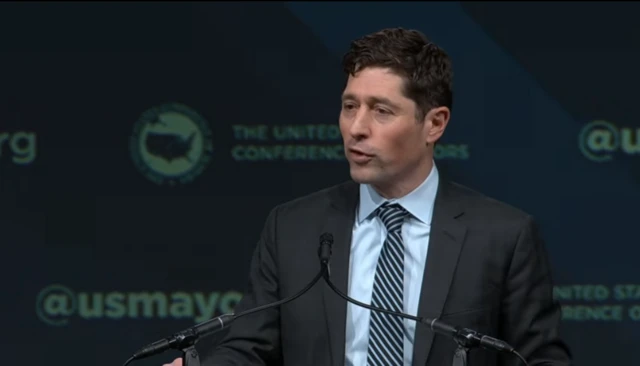 Jacob Frey speaking behind a black lectern, he's in a dark suit, light blue shirt and striped tie