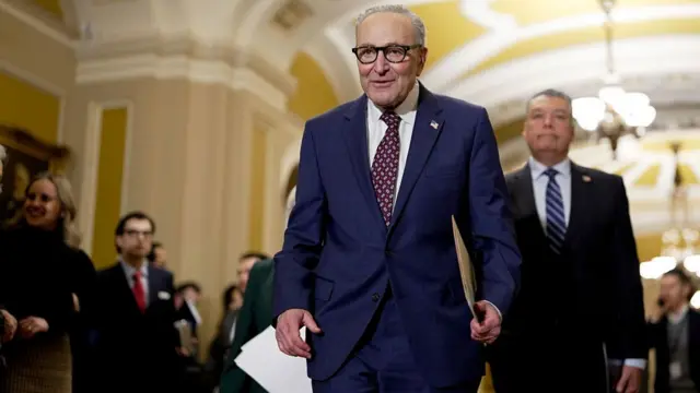 Chuck Schumer walks down the hallway in the Capitol. He wears a dark suit and patterned red tie