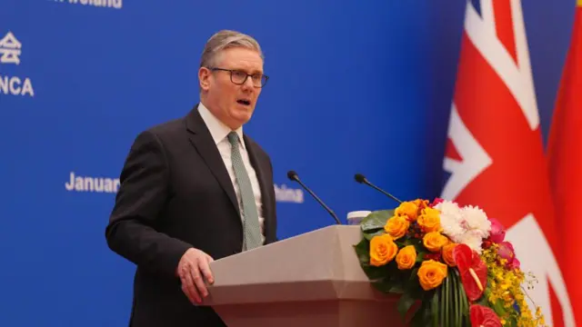 Keir Starmer speaks at a podium with a blue background. A Union Flag can be seen next to him