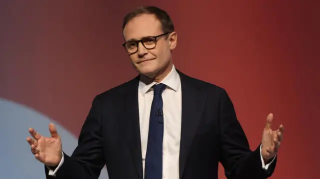 Tom Tugendhat wears a dark suit against a red background