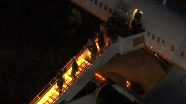 A lit stairwell leading off a plane at night time, several figures are walking down the stairs