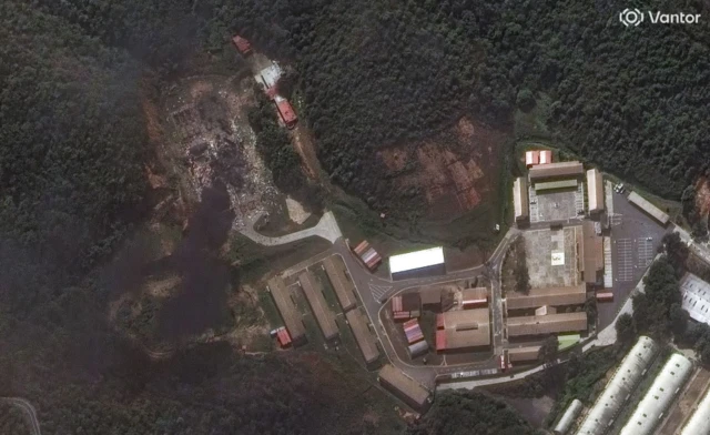 An aerial view of a compound of buildings surrounded by thick forest, there is some black smoke on the left hand side
