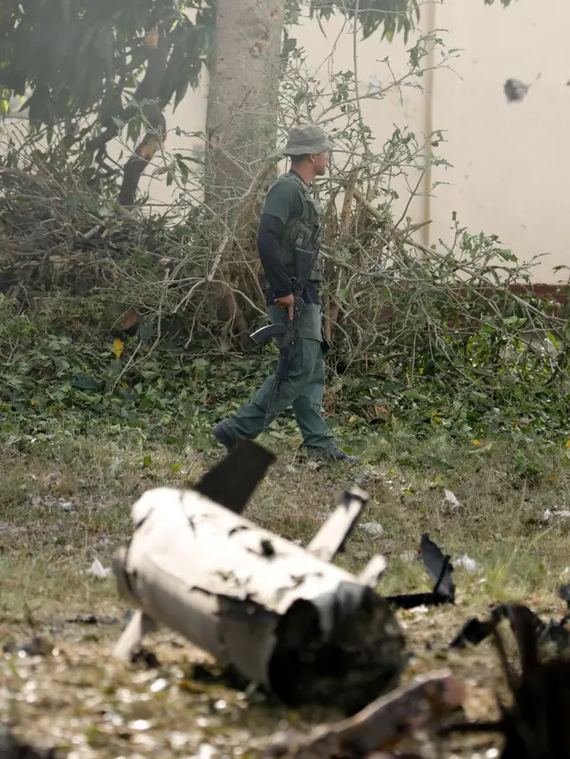 A military personnel member in uniform holding a weapon walks past debris on the ground at La Carlota military air base