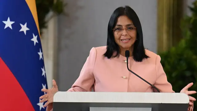 Delcy Rodríguez stands at a podium wearing pink, with her arms held wide