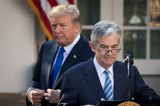 Donald Trump stood behind Jerome Powell who is speaking into a microphone behind a podium.