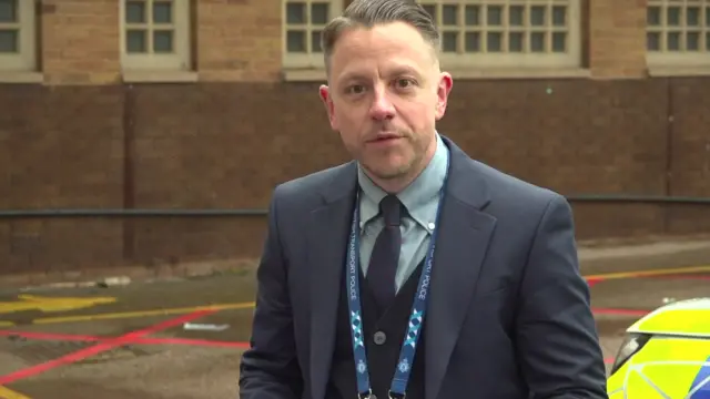 Attwell who wears a suit talks. He stands in an empty car park and you can see the very front of a police car behind him