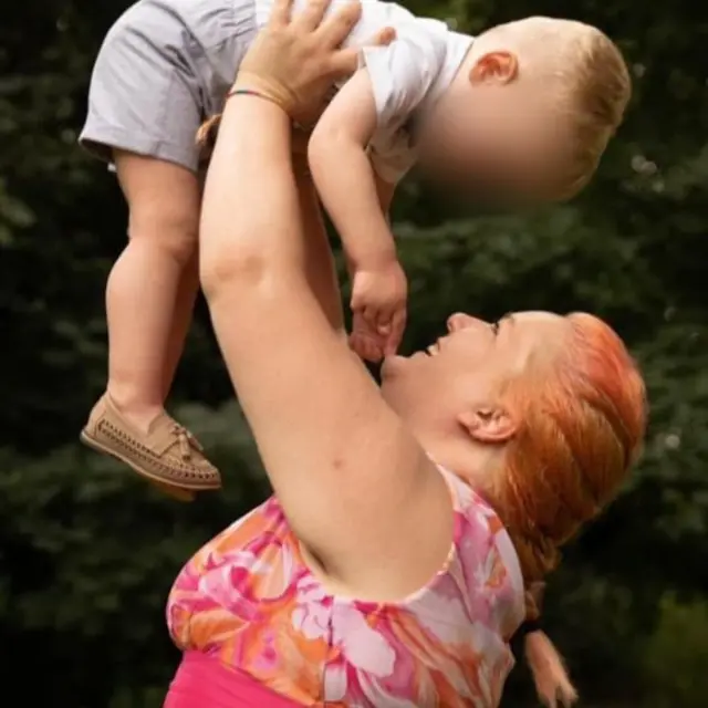 A woman in a pink, orange and white top is smiling and holding a little boy in the air. The woman is smiling and looking into the face of the child. The boy's face is blurred to protect his identity.