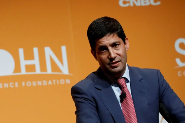 Fed chair nominee Kevin Warsh mid-speech, he's in a dark blue suit, white shirt and white polka dots red tie. Behind him an orange wall with writings in white