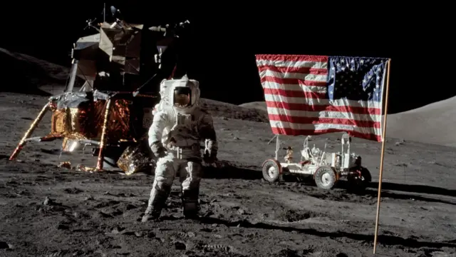 Astronaut Harrison Schmitt, Apollo 17 lunar module pilot, stands near the deployed United States flag on the lunar surface during extravehicular activity (EVA) of NASA's final lunar landing mission in the Apollo series. The Lunar Module (LM) is at left background and the Lunar Roving Vehicle (LRV) at right background (partially obscured).