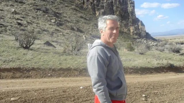 Convicted paedophile Jeffrey Epstein poses in front of a mountain in grey hoodie and red tracksuit trousers
