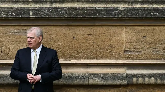 Prince Ansrew in a suit standing against a wall looking to his right