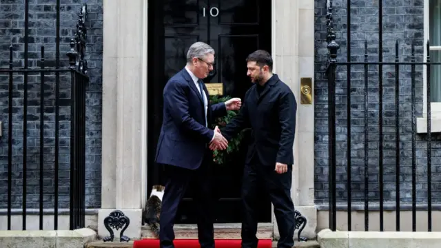 Keir Starmer and Volodymyr Zelensky shaking hands on the steps of 10 Downing Street. The door is behind them, and Larry the cat is sat on the front step.