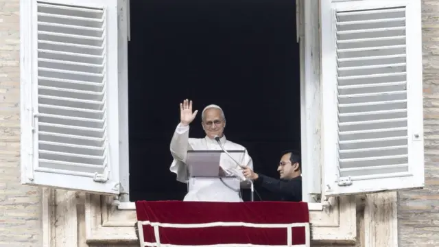 Pope Leo waves through an open window as he stands behind a mic