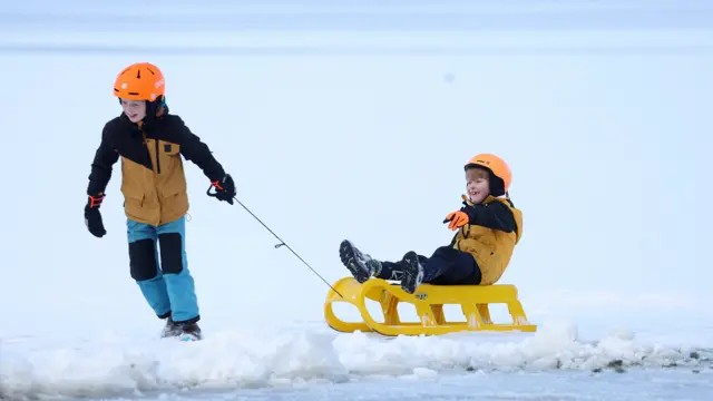 A child tugs another child sitting on a yellow sled along a frozen loch