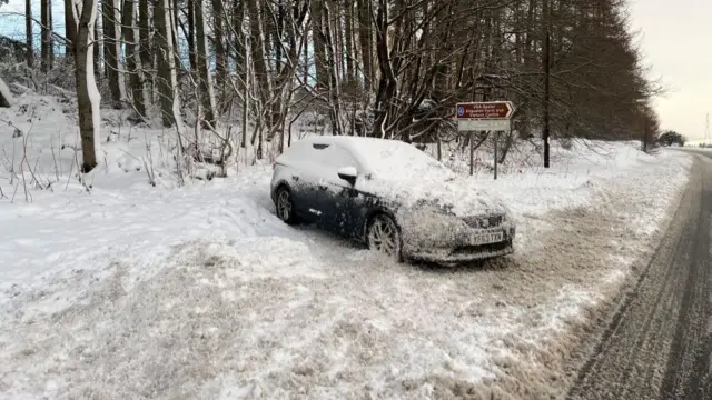 A car covered with snow sits on a snowy verge next to a road covered with melted slush in Aberdeenshire.