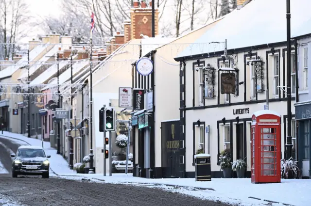 A high street can be seen covered un snow with a car driving along the road