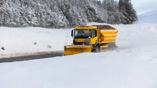 A yellow gritter lorry travels along a cleared road next to mounds of snow