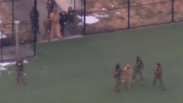 A woman is escorted by three men in uniform, a man is escorted several yards behind her