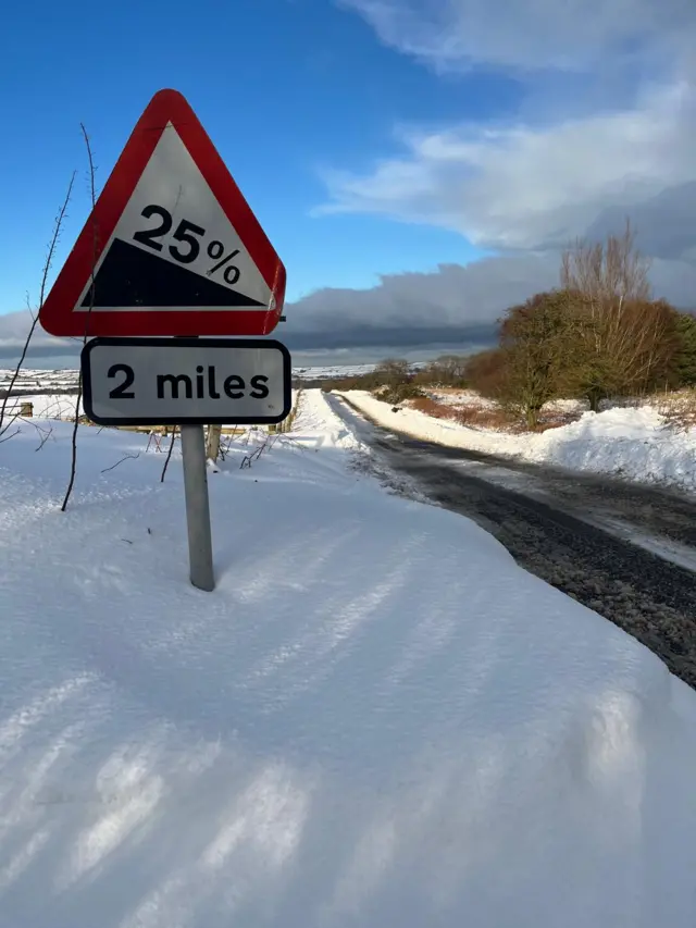 A street sign post partially submerged by snow alongside a rural road in Yorkshire. The road passes through two fields