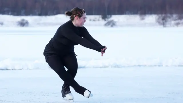 A young woman dressed in black performs manouevres on ice skates on a frozen loch