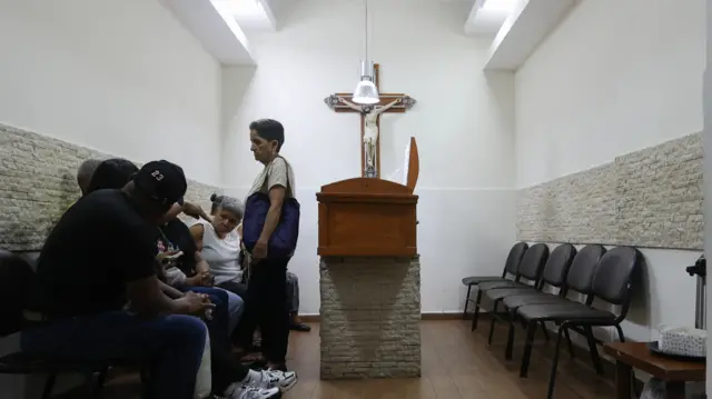 A small, quiet chapel room with several family members sat on one side. An open coffin is in the centre, below a cross with Jesus on. We cannot see inside the coffin.