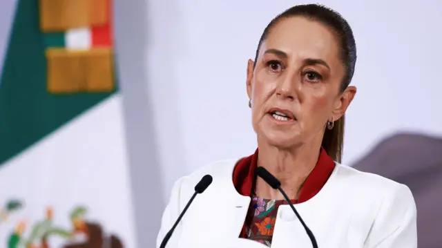 Claudia Sheinbaum. She is a woman with dark-brown hair tied tightly back. She is wearing a white jacket with a red collar and floral patterned top underneath. She is standing at a lectern with two microphones