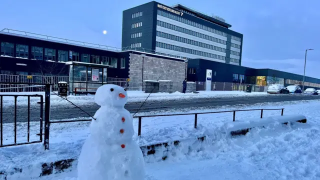 A snowman with twig arms outstretched. It is white and has orange 'buttons' running the length of its body. It has a carrot nose and black eyes. It is standing on a snowy path in front of a black fence and a road which has melted snow on it, In the background is a bus stop and the North East Scotland college building.