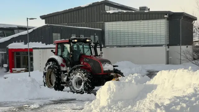 A large red tractor clears a snowdrift outside a dark building with a red doorframe