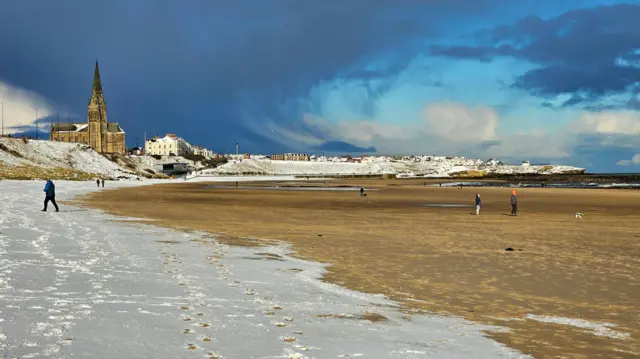 A snowy beach in Tynemouth, a church with a tall bell tower is in the background to the left. There's some people walking along the beach, with two figures to the right observing a dog playing